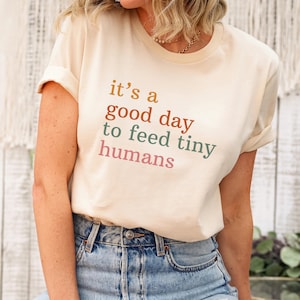Peut inclure: T-shirt crème avec l'inscription "it's a good day to feed tiny humans" en orange, vert et rose. Le t-shirt a des manches retroussées et est associé à un jean bleu. Une petite étiquette "NATURAL" est attachée au t-shirt.