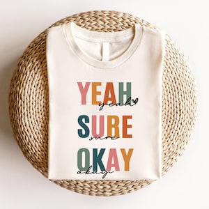 Puede incluir: Una camiseta blanca con el texto "YEAH SURE OKAY" en letras mayúsculas de colores. Las palabras son de diferentes colores, incluyendo rosa, naranja, verde y azul.