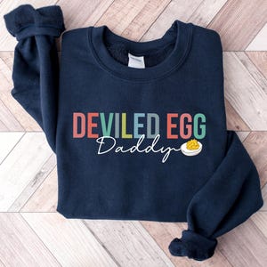 May include: Navy blue sweatshirt with the words "DEVILED EGG Daddy" in colorful letters. A small graphic of a deviled egg is below the text. The sweatshirt has a crew neck and long sleeves.