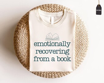 May include: Cream-coloured t-shirt with the text "emotionally recovering from a book" in teal. The shirt features a simple illustration of an open book above the text. The shirt is displayed on a woven, circular surface.