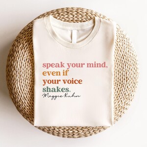 Può includere: T-shirt color crema con la citazione "speak your mind, even if your voice shakes." in lettere rosa, arancioni e verdi. La maglietta presenta anche la firma "Maggie Kuhn" in nero. È appoggiata su una superficie circolare intrecciata.