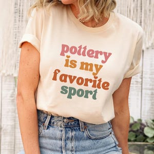 May include: A cream-coloured t-shirt with the text "pottery is my favourite sport" in pink, orange, brown, and green. The shirt has rolled-up sleeves and is worn with blue denim jeans. A tag is attached to the shirt.