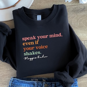 Può includere: Felpa nera con la citazione "speak your mind, even if your voice shakes" in arancione, rosa e verde, attribuita a Maggie Kuhn. La felpa è abbinata a jeans blu e un cappello bianco.