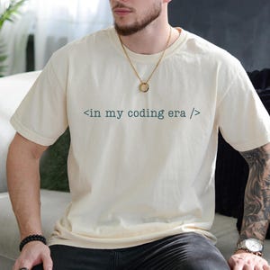 May include: A person wearing a cream coloured t-shirt with the text "< in my coding era />" printed on the front. The person is wearing a gold chain necklace.