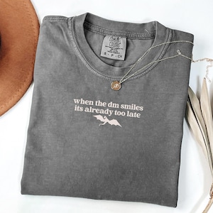May include: A gray Comfort Colors t-shirt with the text "when the dm smiles its already too late" and a dragon graphic. A gold necklace with a pendant rests on the shirt. The shirt is folded on a white surface.