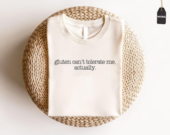 Könnte beinhalten: Cremefarbenes T-Shirt mit dem schwarzen Aufdruck: "gluten can't tolerate me, actually". Das Shirt liegt auf einer runden, geflochtenen Unterlage. Ein kleines, schwarzes Etikett mit dem Wort "NATURAL" ist angebracht.
