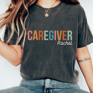 May include: Dark gray t-shirt with the word "CAREGIVER" in colorful block letters and the name "Rachel" in white script. The shirt is a casual style, suitable for everyday wear.