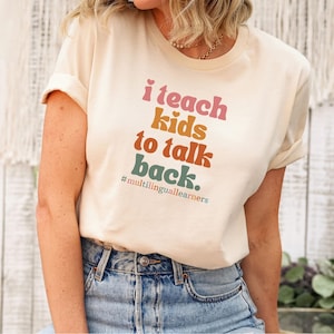 May include: A cream-coloured t-shirt with the text "I teach kids to talk back." in pink, orange, and green. The hashtag #multilinguallearners is below the text. The shirt is worn with blue jeans.