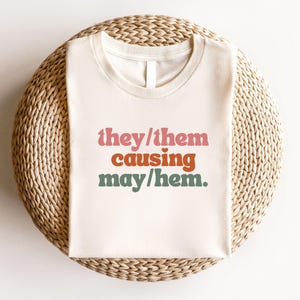 May include: A cream-coloured t-shirt featuring the text "they/them causing may/hem" in pink, orange, and green. The shirt is placed on a woven, circular surface, with a small tag that says "natural".