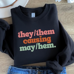 May include: Black crew neck sweatshirt featuring the text "they/them causing may/hem" in pink, orange, and green. The sweatshirt is folded with blue jeans and a white hat in the background. The sweatshirt is made of a soft material.