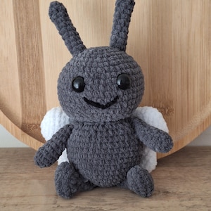 May include: A handmade gray crochet insect toy with large black eyes, a smiling mouth, and white wings. The plush toy has long, upright antennae and is sitting on a wooden surface. This soft toy is perfect for children.