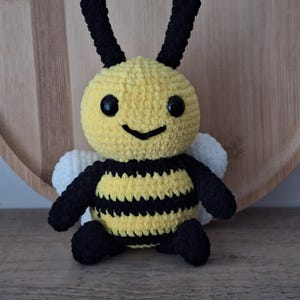 May include: A handmade crochet bee plushie with a yellow body, black stripes, and black antennae. The bee has black eyes and a smiling mouth. It has white wings and black legs and arms. The plushie is sitting on a wooden surface.