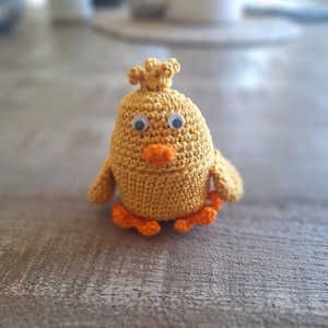 May include: A crocheted yellow chick with orange feet and white eyes. The chick is sitting on a wooden surface.