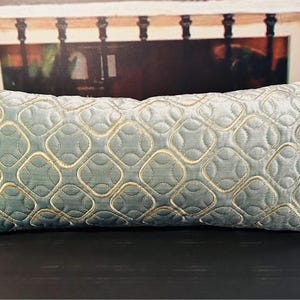 May include: A rectangular decorative pillow in a light blue hue, featuring a quilted pattern of interlocking circles outlined in a lighter gold color. The pillow has a textured surface and a decorative trim along the edges.