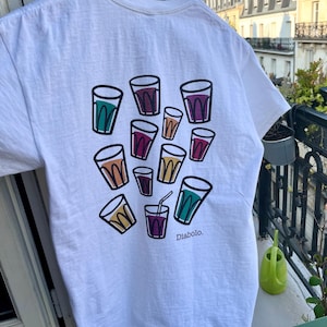 May include: White t-shirt featuring a colorful graphic of various drink glasses with black outlines. The glasses are filled with different colored liquids. The word "Diabolo" is printed in black below the glasses. The shirt is hanging on a wooden hanger.