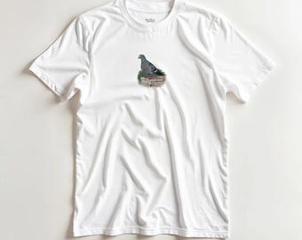 May include: White t-shirt featuring a graphic of a pigeon perched on a rock. The pigeon is depicted in shades of gray, green, and brown. The shirt has a classic crew neck design and short sleeves. The shirt is made of a soft, lightweight fabric.