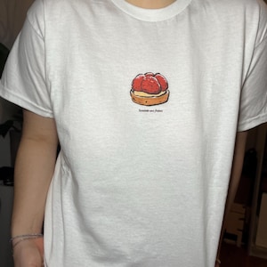 May include: White t-shirt featuring a small illustration of a strawberry tart. The graphic shows a pastry base topped with red strawberries. The text "Tartelette aux Fraises" is printed below the tart.