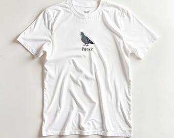 May include: White t-shirt featuring a detailed illustration of a pigeon with gray, black, and red coloring. The word "Paris" is written in a script font below the bird. The shirt has a crew neck and short sleeves.