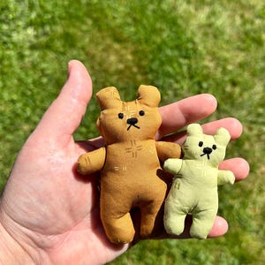 Op de afbeelding: Twee kleine knuffelberen gemaakt van stof. De grotere beer is bruin met geborduurde gelaatstrekken en subtiele kruissteekdetails op de borst. De kleinere beer is lichtgroen met vergelijkbare geborduurde gelaatstrekken.