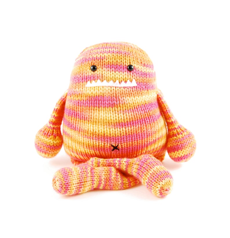 Pode incluir: Um brinquedo de pel&uacute;cia de malha na forma de um monstro com um padr&atilde;o listrado rosa e laranja. O brinquedo tem uma boca branca com dentes e olhos pretos.