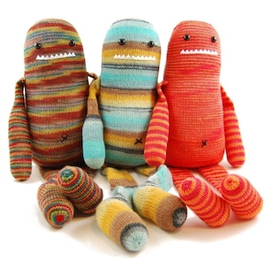 Puede incluir: Tres juguetes de peluche tejidos con patrones a rayas y sonrisas con dientes. Los juguetes son rojo, azul y marrón con rayas amarillas y naranjas.