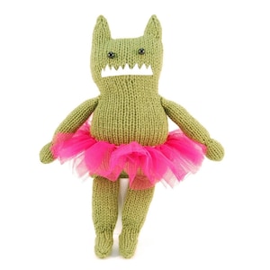 Puede incluir: Un juguete monstruo de punto verde que lleva un tutú de tul rosa. El monstruo tiene una sonrisa de dientes y ojos negros.