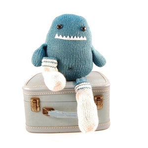 May include: A blue knitted plush toy with a toothy grin sits on a vintage blue suitcase. The toy has white socks with blue stripes.