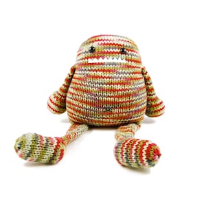 Puede incluir: Un juguete de peluche de punto a rayas con una sonrisa de dientes y ojos grandes. El juguete está sentado con las piernas extendidas.