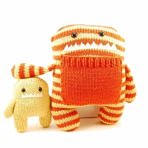 May include: Two knitted plush toys, one orange and yellow striped with a white toothy grin, and one yellow with a white toothy grin. The larger toy is standing and the smaller toy is sitting.