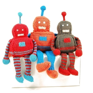 May include: Three knitted robot toys, one red, one orange, and one grey, sitting on a white surface. Each robot has a different coloured antenna and striped legs.