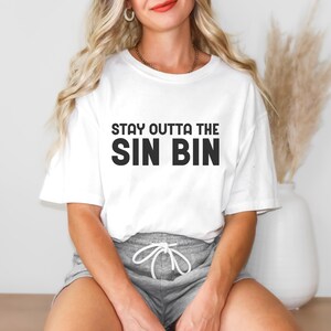 May include: White t-shirt with the text "STAY OUTTA THE SIN BIN" in black block letters. The shirt is worn with gray shorts. The overall style is casual and comfortable.