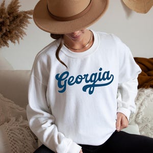 Op de afbeelding: Witte sweatshirt met het woord "Georgia" in een donkerblauwe script. De sweatshirt heeft lange mouwen en een ronde hals. De afbeelding toont een persoon die de sweatshirt draagt.