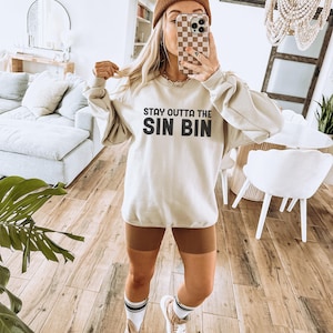 May include: Cream-colored sweatshirt with the text "STAY OUTTA THE SIN BIN" in black. The person is wearing brown shorts, a brown beanie, and white sneakers with brown accents. The person is holding a phone with a checkered case.