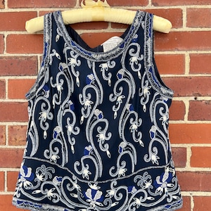 K&ouml;nnte beinhalten: Marineblaues &auml;rmelloses Oberteil mit wei&szlig;er und blauer Blumenstickerei. Das Oberteil h&auml;ngt an einem Vintage-Holzb&uuml;gel vor einer roten Backsteinmauer. Das florale Design ist aufwendig und detailliert.
