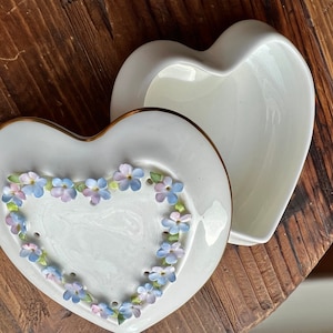 May include: White porcelain heart-shaped trinket box with a gold trim and a floral design of blue and pink forget-me-nots.