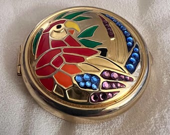 May include: A round, gold-toned compact with a colorful parrot design. The parrot features red, orange, and yellow details, with green leaves and blue and purple accents. The compact is closed and sits on a white surface.