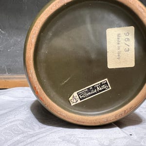 May include: Close-up of the bottom of a dark green ceramic vase with a brown rim. The vase has two stickers, one with the text "Made in Italy" and the other with the text "Created in Italy for Rosenthal Netter".