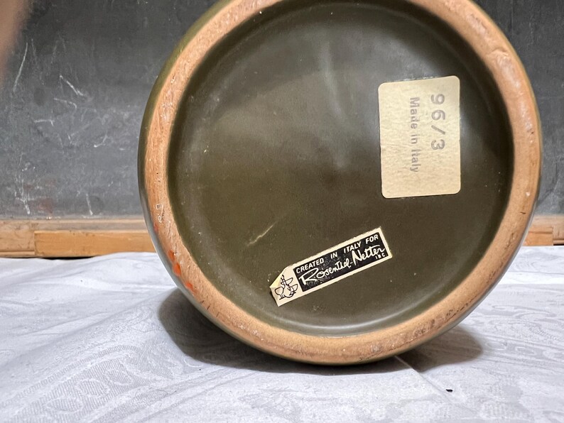 May include: Close-up of the bottom of a dark green ceramic vase with a brown rim. The vase has two stickers, one with the text "Made in Italy" and the other with the text "Created in Italy for Rosenthal Netter".