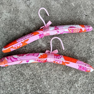 May include: Two padded clothes hangers with a pink, orange, and white floral pattern. Each hanger has a pink metal hook and a matching pink ribbon tied at the neck. The hangers are designed for clothing storage.