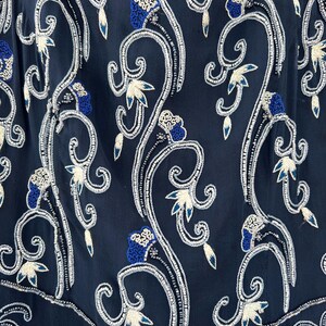 K&ouml;nnte beinhalten: Nahaufnahme eines marineblauen Kleidungsst&uuml;cks mit aufwendiger Blumenstickerei. Das Design zeigt wei&szlig; und blau gestickte Blumen und wirbelnde Ranken, akzentuiert mit silbernen Perlen. Das Gesamtmuster ist symmetrisch und detailliert.