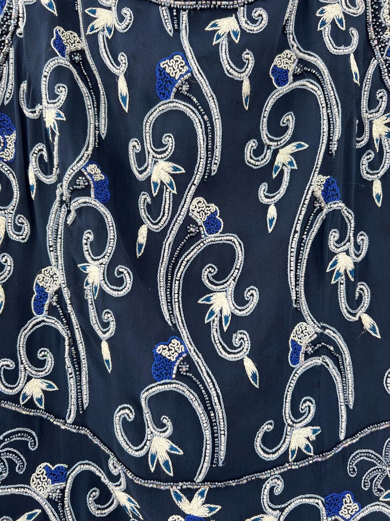 K&ouml;nnte beinhalten: Nahaufnahme eines marineblauen Kleidungsst&uuml;cks mit aufwendiger Blumenstickerei. Das Design zeigt wei&szlig; und blau gestickte Blumen und wirbelnde Ranken, akzentuiert mit silbernen Perlen. Das Gesamtmuster ist symmetrisch und detailliert.