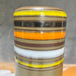 May include: A cylindrical ceramic vase with horizontal bands of color. The bands include yellow, black, orange, brown, white, and olive green. The vase has a glossy finish and a rounded top.