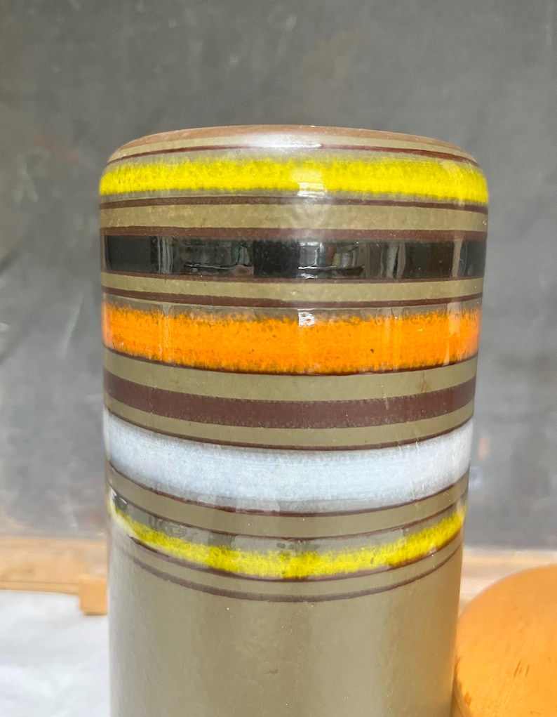 May include: A cylindrical ceramic vase with horizontal bands of color. The bands include yellow, black, orange, brown, white, and olive green. The vase has a glossy finish and a rounded top.
