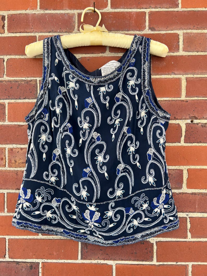 K&ouml;nnte beinhalten: Marineblaues &auml;rmelloses Oberteil mit wei&szlig;er und blauer Blumenstickerei. Das Oberteil h&auml;ngt an einem Vintage-Holzb&uuml;gel vor einer roten Backsteinmauer. Das florale Design ist aufwendig und detailliert.