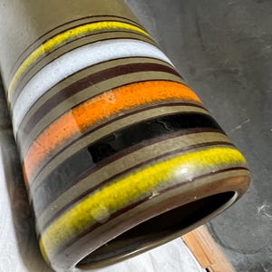 May include: A tall, olive-green ceramic vase with horizontal bands of color. The bands include yellow, white, orange, black, and brown. The vase has a wide opening and a rounded base. The design is a retro style.