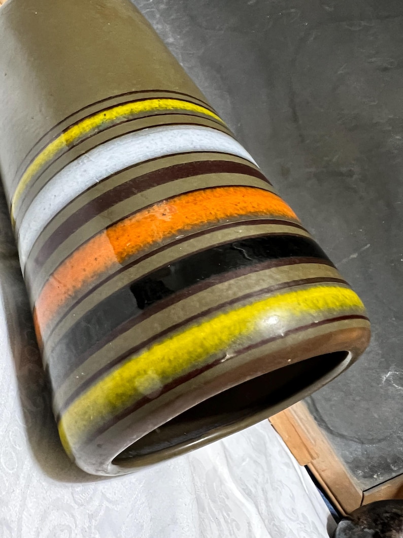 May include: A tall, olive-green ceramic vase with horizontal bands of color. The bands include yellow, white, orange, black, and brown. The vase has a wide opening and a rounded base. The design is a retro style.