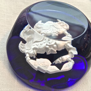 Pu&ograve; includere: Un fermacarte in vetro blu scuro con una scultura bianca e dettagliata di un granchio all'interno. Il granchio &egrave; il punto focale, con dettagli intricati. Il fermacarte ha una forma arrotondata e riflette la luce.