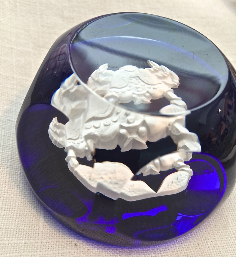 Pu&ograve; includere: Un fermacarte in vetro blu scuro con una scultura bianca e dettagliata di un granchio all'interno. Il granchio &egrave; il punto focale, con dettagli intricati. Il fermacarte ha una forma arrotondata e riflette la luce.
