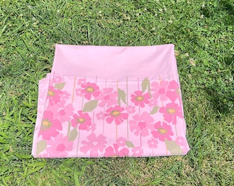May include: A pink floral bed sheet set on green grass. The sheet features a repeating pattern of pink flowers with yellow centers and green leaves. The top portion of the sheet is a solid pink color.
