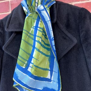 May include: A colorful scarf with a blue, green, and white geometric pattern is draped over a black coat. The scarf's design features intersecting lines and rectangular shapes. The coat has a classic, tailored design.